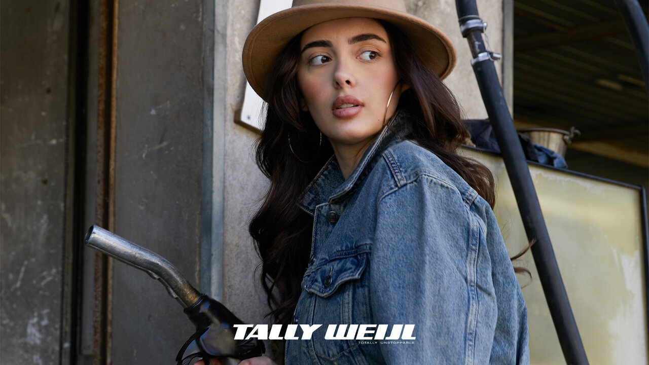 Eine junge Frau in Jeansjacke und Hut, die eine Zapfpistole in der Hand hält, steht vor einer Betonwand mit einem Metallrohr und einem Schild mit der Aufschrift "TALLY WELL".