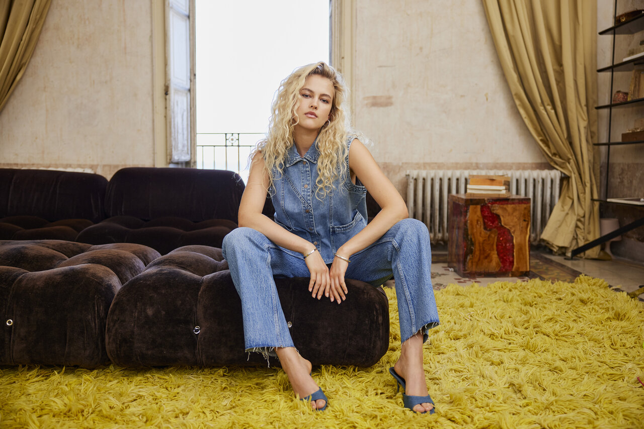 Eine Frau in einem Jeansoverall und blauen Stöckelschuhen sitzt auf einer braunen Couch und blickt in die Kamera, unter ihren Füßen liegt ein gelber Zottelteppich.