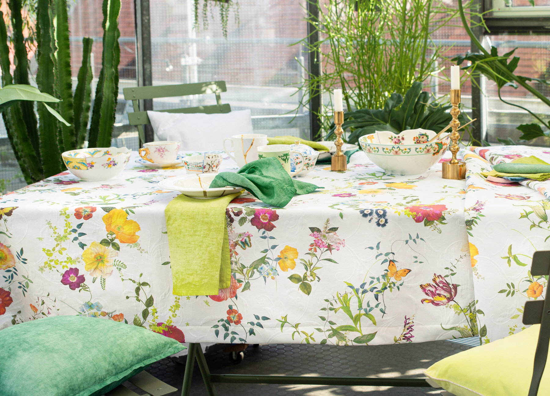 Ein weißes Tischtuch mit Blumenmuster wird mit einer grünen Serviette, goldenen Kerzenhaltern und einer weißen Schale gedeckt, dazu ein grüner Stuhl und ein großes Fenster mit einem grünen Vorhang.