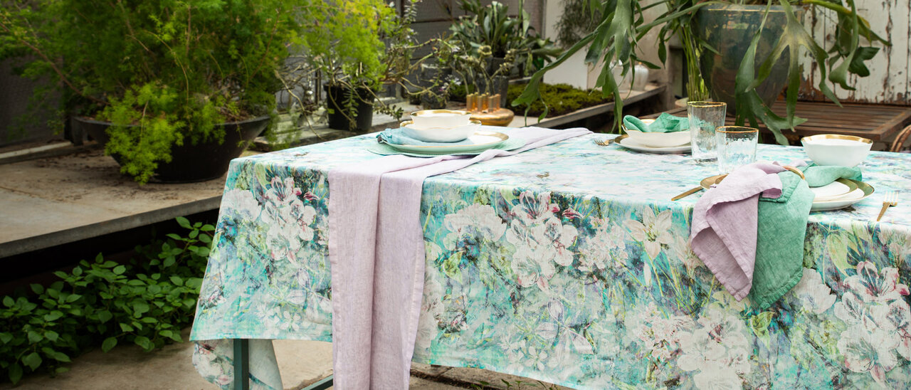 Der Tisch für zwei Personen auf dem Balkon ist mit einer blau-weißen Blumentischdecke, weißen Tellern und Silberbesteck gedeckt, daneben stehen eine Vase mit Grünzeug und eine Topfpflanze.