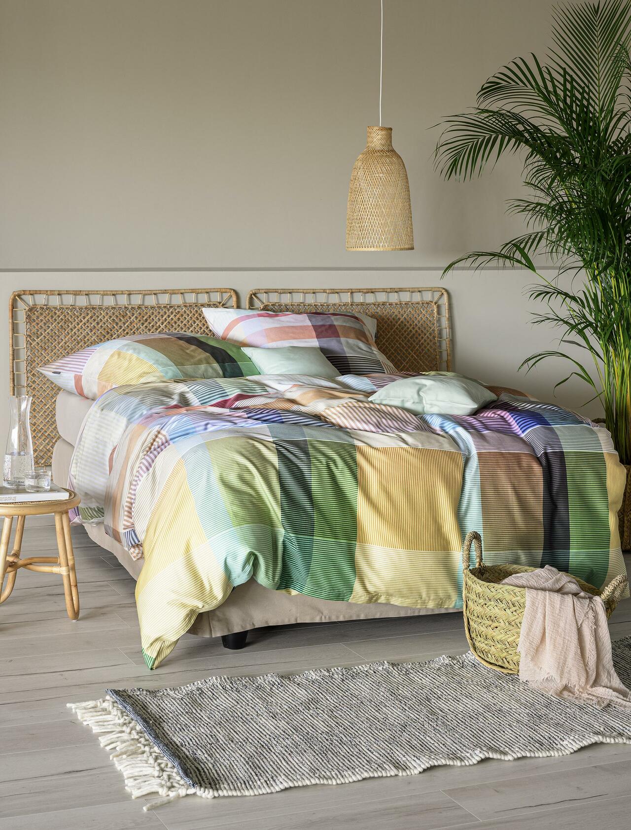 In einem gemütlichen Schlafzimmer stehen ein Bett mit einer lebhaft karierten Bettdecke, ein geflochtenes Kopfteil, eine Hängeleuchte, ein geflochtener Korb und ein kleiner Beistelltisch aus Holz.