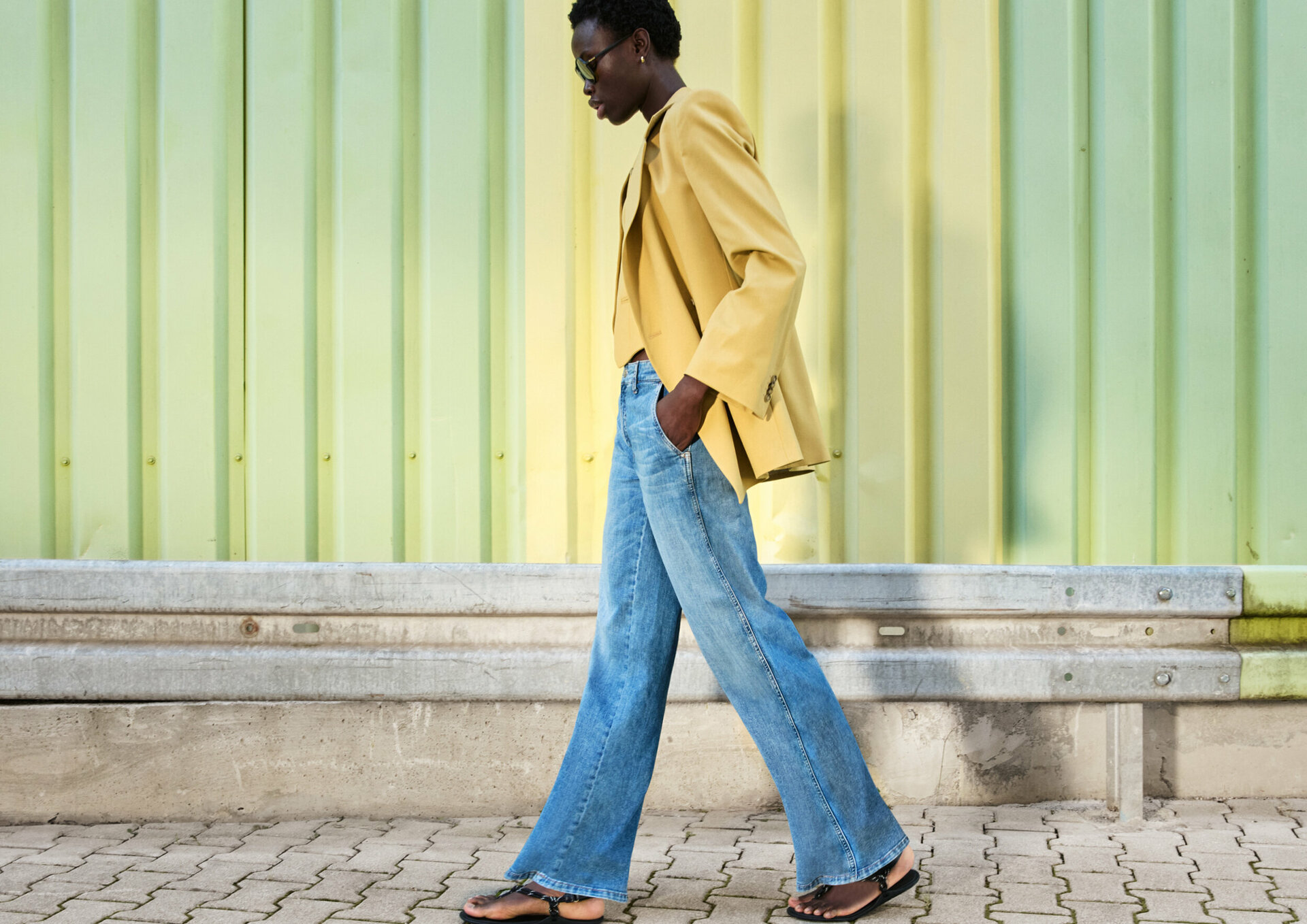 Eine Person in einem gelben Blazer, blauen Jeans und schwarzen Sandalen geht auf einem gemauerten Bürgersteig, mit einer grünen Wellblechwand im Hintergrund.