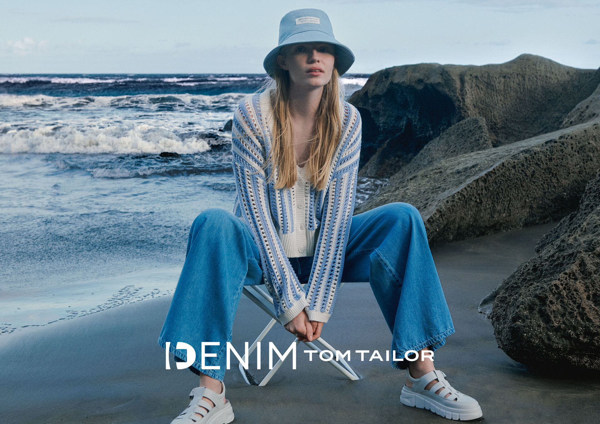 Eine Frau in einem blau-weiß gestreiften Pullover, blauen Jeans und weißen Turnschuhen sitzt auf einem Strandstuhl und blickt auf das Meer. In der Ecke ist das IDENIM TOM TAILOR-Logo zu sehen.