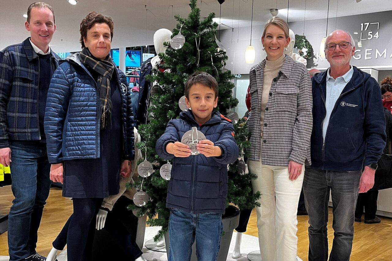 Ein kleiner Junge in einer blauen Jacke hält ein silbernes Ornament, umgeben von einer sechsköpfigen Familie in einem modernen Geschäft mit Weihnachtsbaum und Schaufensterpuppen.