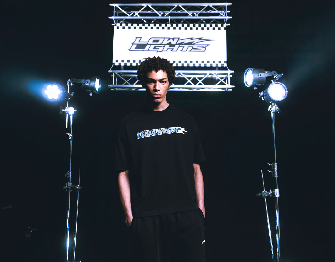 Ein junger Mann in einem schwarzen T-Shirt mit "Low Lights"-Logo steht vor einem schwarz-weiß-karierten Hintergrund, flankiert von zwei Scheinwerfern.